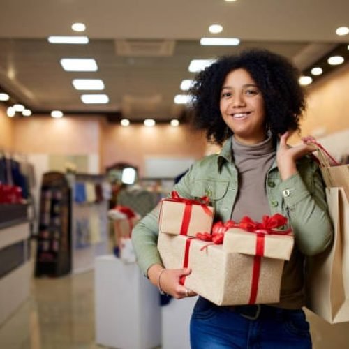 Happy african american girl shopping gifts in mall on christmas sale. New year holidays shopping idea concept. Smiling mixed race woman with colorful paper gift boxes wearing christmas hat.